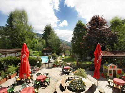 Garten mit Tischen, roten Sonnenschirmen, Bäumen und Bergen im Hintergrund an einem sonnigen Tag.