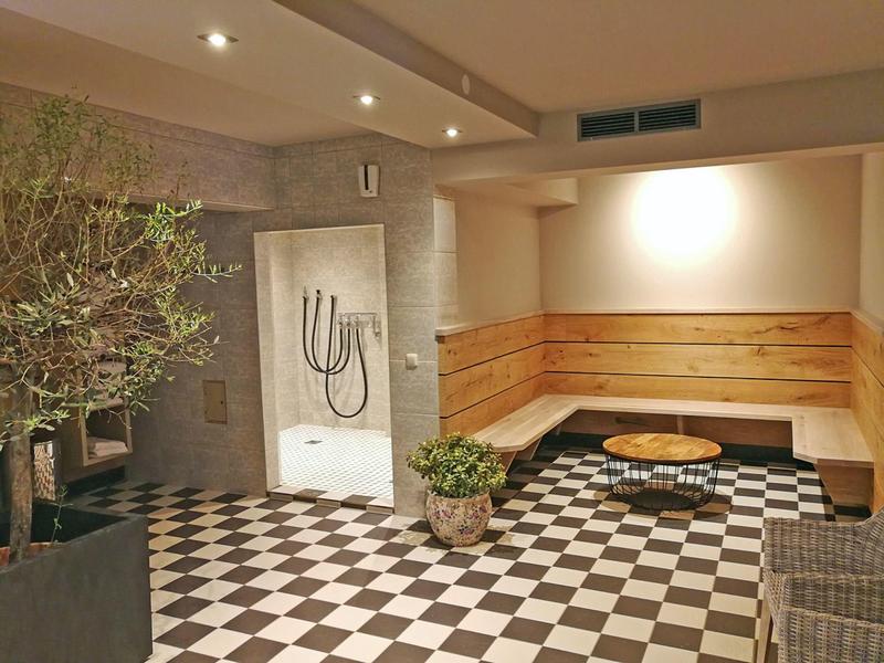 Gezellige hotel lounge met zwart-wit geblokte vloer, houten bank, ronde tafel en planten onder zachte verlichting.