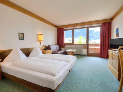 Bright hotel room with double bed, seating area, TV, and balcony with mountain view.