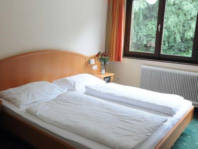 Bright hotel room with double bed, wooden frame, bedside table, and window overlooking greenery.