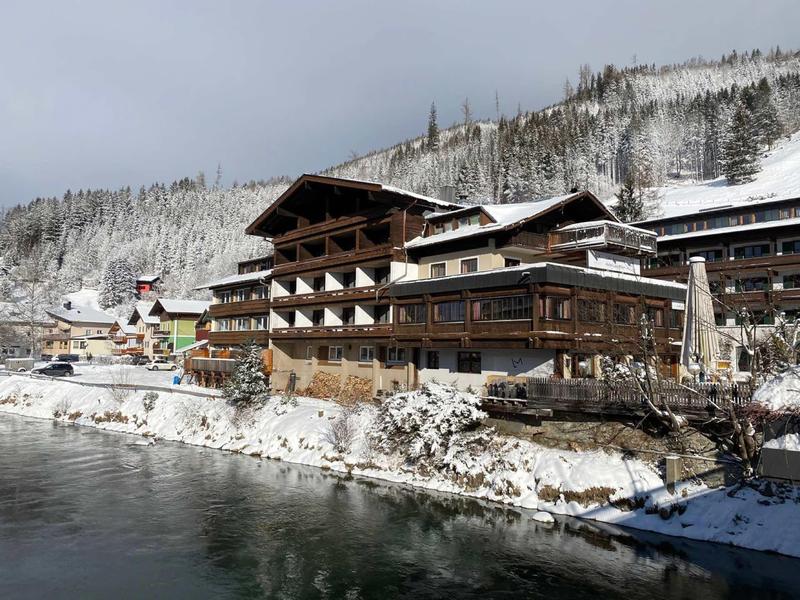 Snow-covered hotel building on the shore of a calm lake in a mountain landscape.