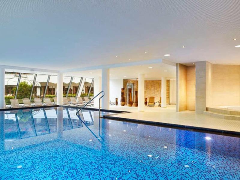 Piscina interna luminosa con grandi finestre che si affacciano su un giardino con lettini.