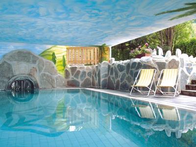 Piscina coperta con arco decorativo in pietra, sedie a sdraio e soffitto dipinto a cielo.