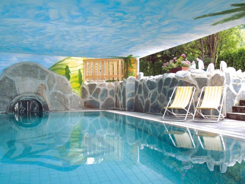 Piscina coperta con arco decorativo in pietra, sedie a sdraio e soffitto dipinto a cielo.