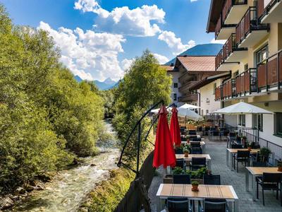 Terrasse eines Hotels am Fluss mit Tischen, Stühlen und roten Sonnenschirmen neben Bäumen.