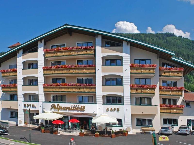 Wielopiętrowy budynek hotelu z balkonami i tarasem kawiarni na tle zalesionych gór.