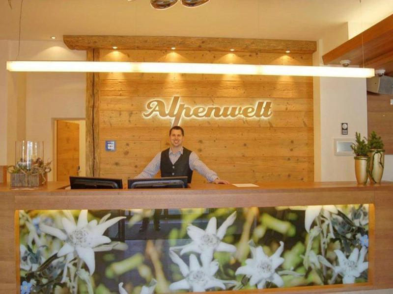 Recepcja z drewnianą okładziną i obrazem kwiatów, pracownik za ladą w hotelu Alpenwelt.