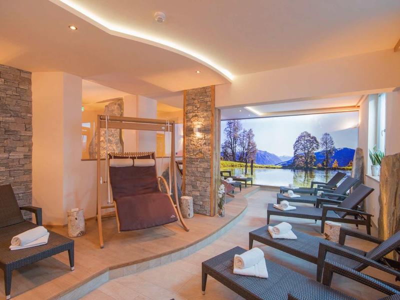Pokój relaksacyjny z leżakami, drewnianym fotelem i widokiem na teren zewnętrzny z jeziorem i górami.