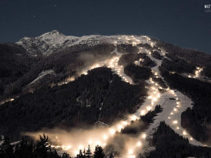 Oświetlone stoki narciarskie na górze w nocy z zaśnieżonym szczytem i ciemnym niebem.