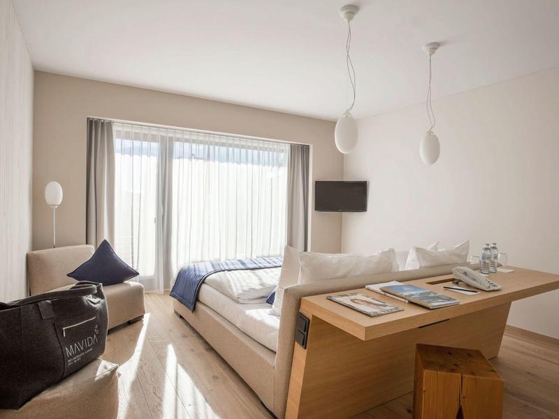 Habitación de hotel luminosa con cama doble, sillones y ventana grande con cortinas.