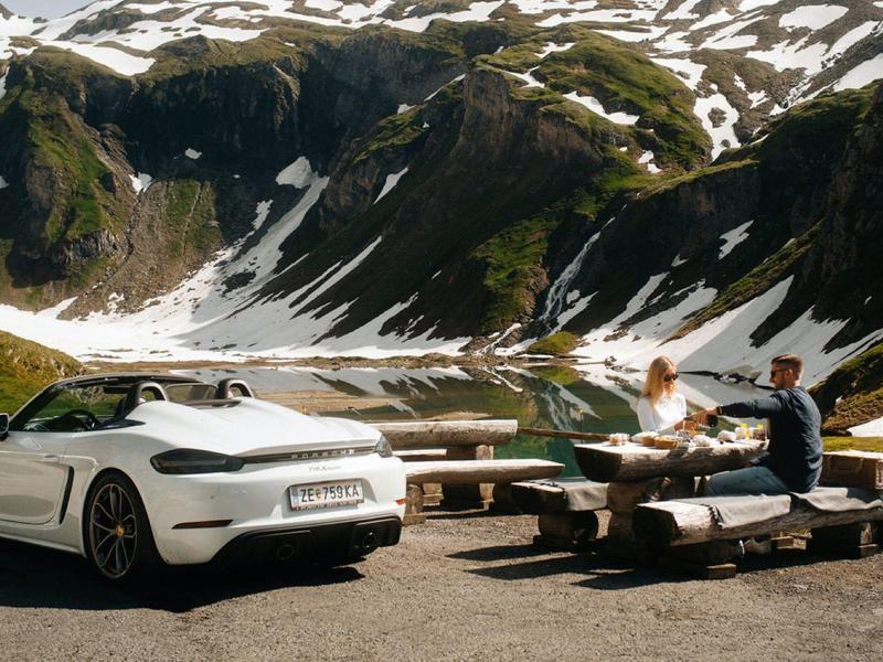 Coche deportivo blanco junto a dos personas en mesa de madera en montañas con nieve.