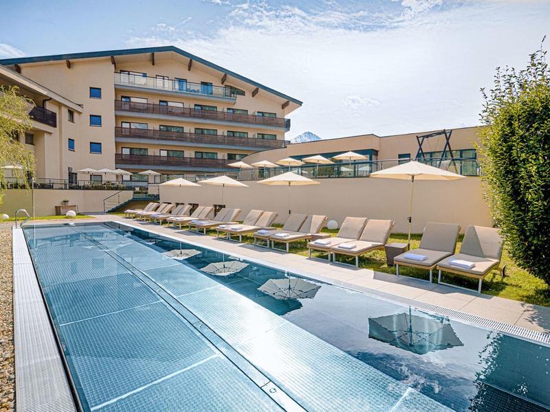Piscina al aire libre con tumbonas y sombrillas frente a un hotel moderno bajo un cielo azul.