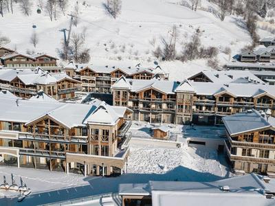 Schneebedeckte Alpenchalets aus Holz mit großen Balkonen vor einem bewaldeten Berghang.