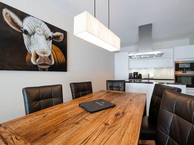 Modern dining room with wooden table, black chairs, cow painting, and sleek kitchen in background.