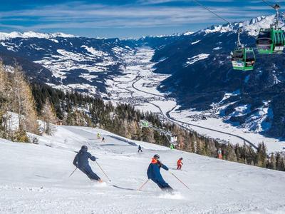Two skiers descend a snowy mountain with a valley and chairlift in the background.