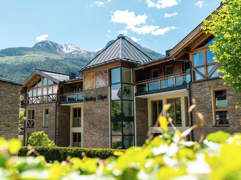 Hotel building with glass section, surrounded by mountains and greenery under a blue sky.