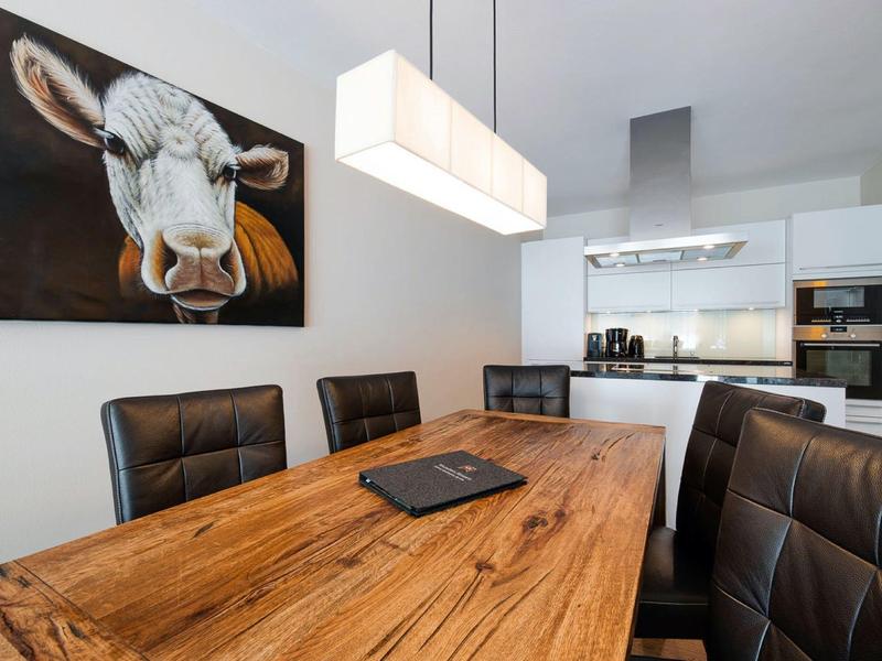 Modern dining room with wooden table, black chairs, cow painting, and sleek kitchen in background.