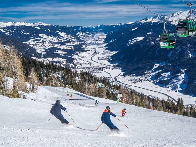 Two skiers descend a snowy mountain with a valley and chairlift in the background.
