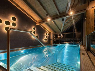 Modernes Hallenbad mit blau beleuchtetem Wasser, Holzdecke und rundlichen Wandlampen.