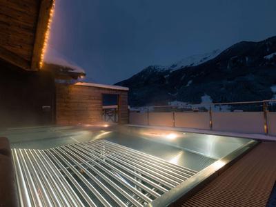 Beleuchteter Außenpool auf Holzboden bei Nacht mit Bergkulisse und Holzhaus im Hintergrund.