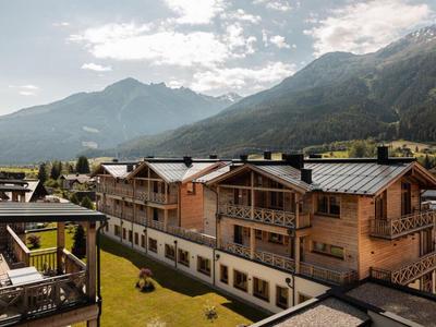 Holzhäuser in Berglandschaft mit grünen Wiesen unter wolkigem Himmel und hohen Bergen im Hintergrund.