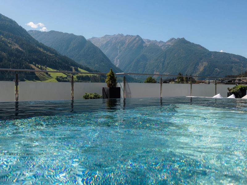 Blauer Pool mit klarem Wasser vor Glasgeländer, dahinter grüne Berge und blauer Himmel.