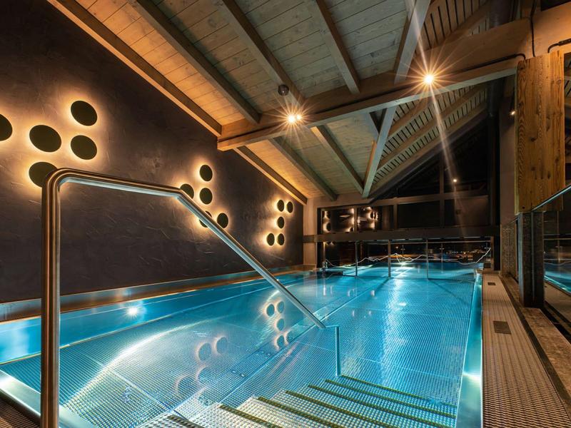 Modernes Hallenbad mit blau beleuchtetem Wasser, Holzdecke und rundlichen Wandlampen.