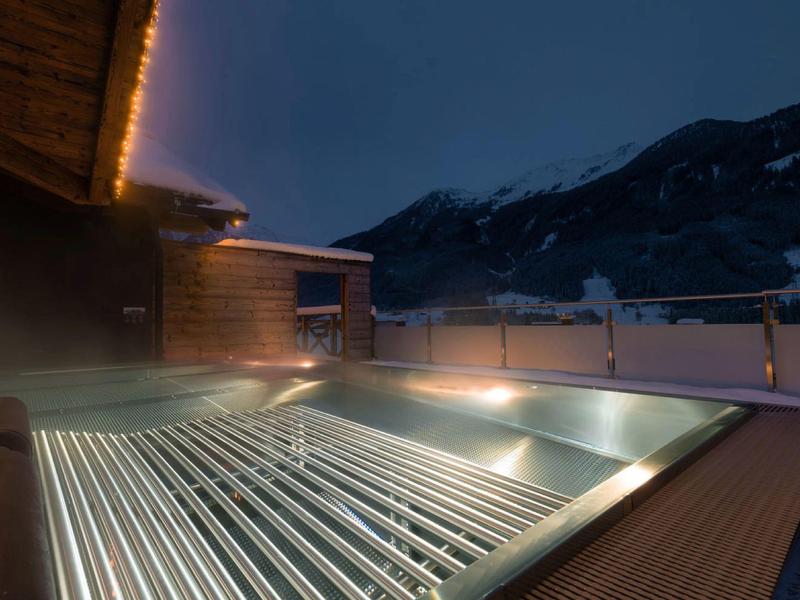 Beleuchteter Außenpool auf Holzboden bei Nacht mit Bergkulisse und Holzhaus im Hintergrund.