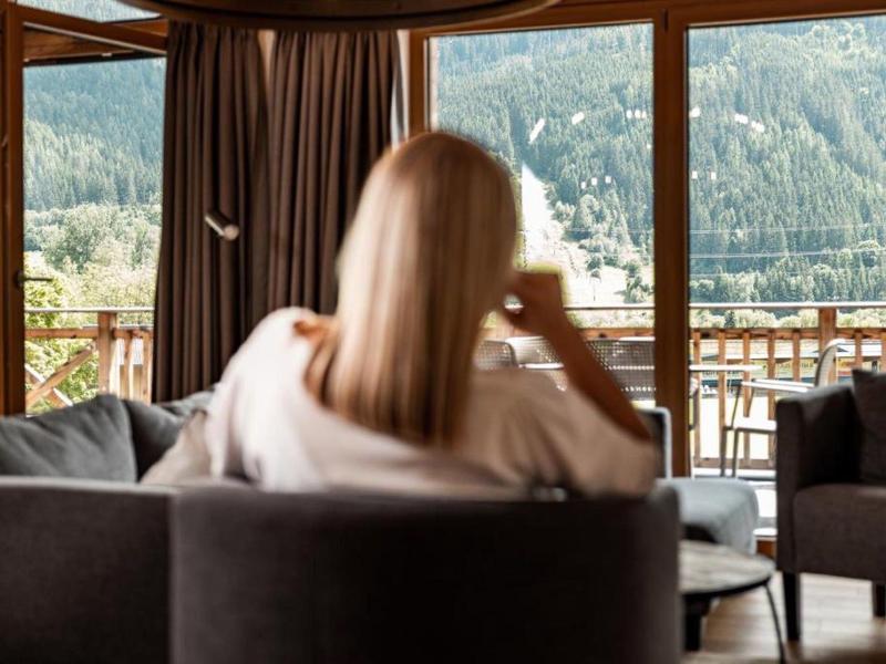 Frau mit blonden Haaren sitzt entspannt in modernem Wohnzimmer mit Ausblick auf grüne Berge.