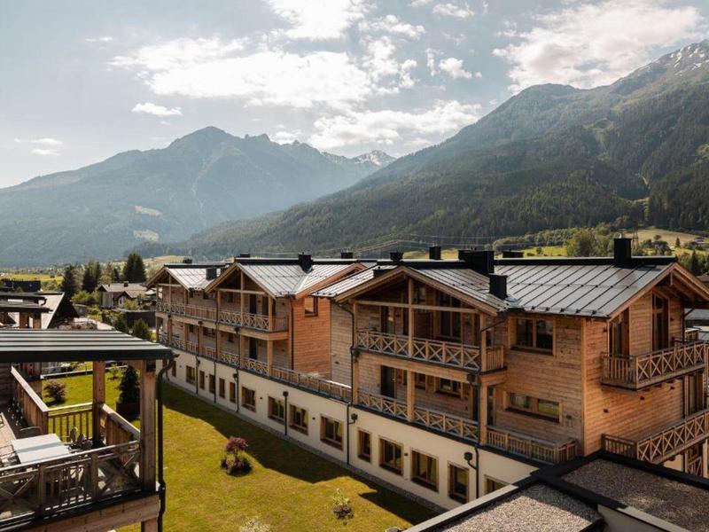Holzhäuser in Berglandschaft mit grünen Wiesen unter wolkigem Himmel und hohen Bergen im Hintergrund.