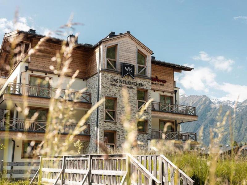 Mehrstöckiges Haus mit Balkonen vor Bergkulisse, umgeben von Gras und Holzzaun bei klarem Himmel.