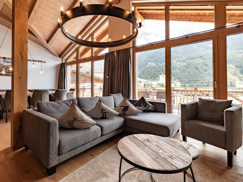 Wohnzimmer mit grauer Polstergarnitur, Holzdecke, großem Fenster und Bergblick.