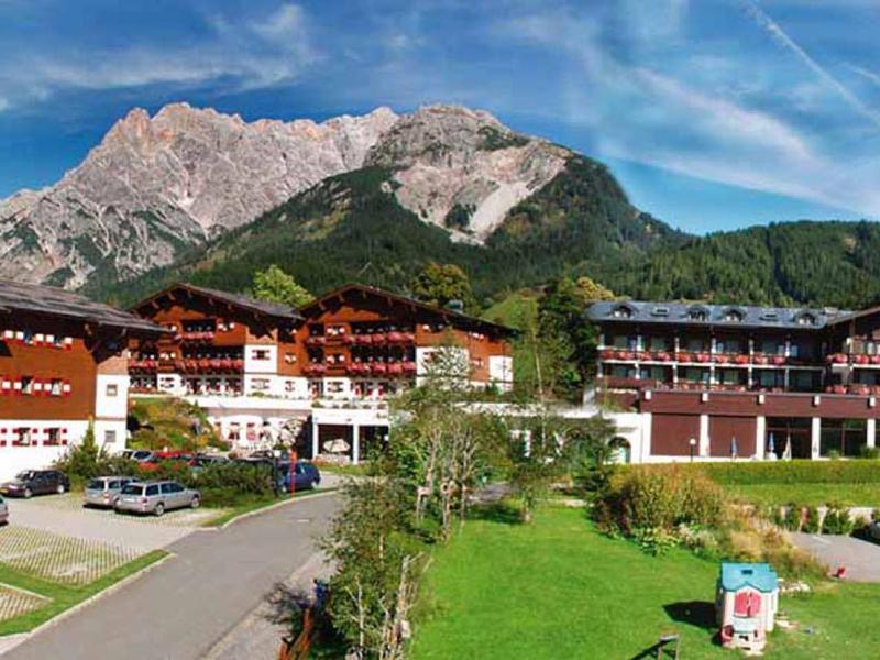 Velký hotel s několika budovami před zalesněnými horami pod jasnou oblohou.