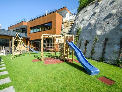Modern playground with slide, climbing frame, and swing next to a contemporary building and rock wall.