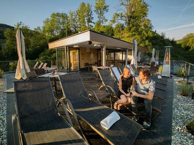 Moderne Terrasse mit Liegestühlen, zwei Personen sitzen und unterhalten sich bei Sonnenschein.