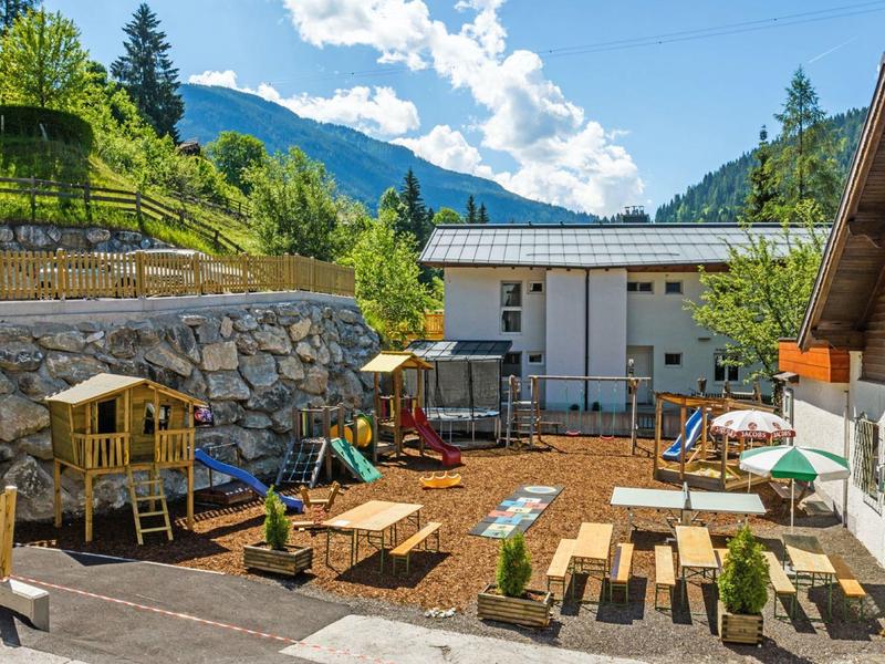 Outdoor area with tables, chairs, and a playground in front of mountains and houses.