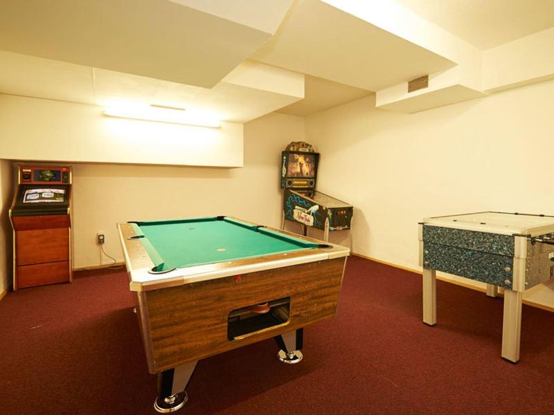 A bright game room with a pool table, pinball machine, and foosball on red carpet.