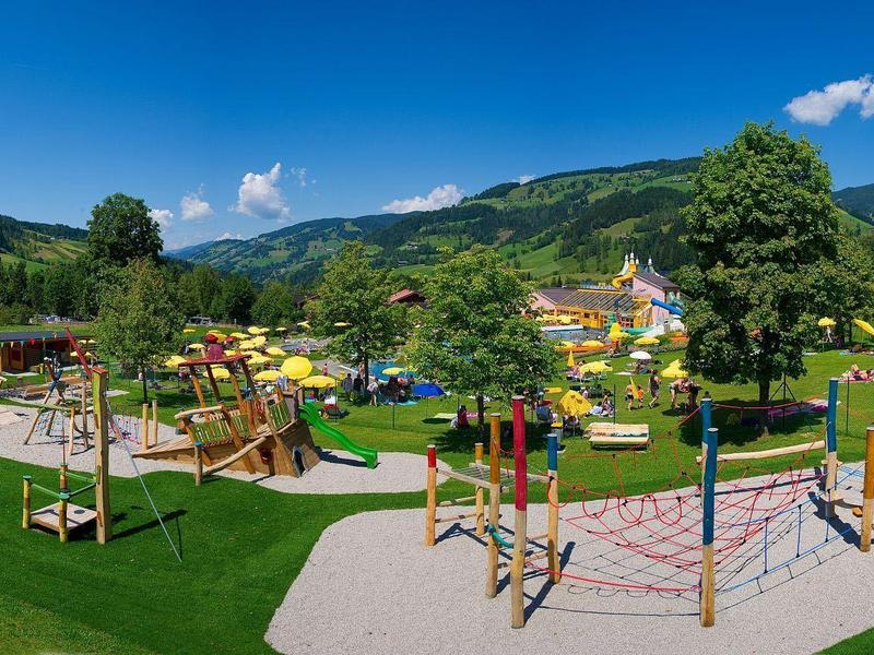 Large children's playground with various colorful play equipment in a green park set against forested hills.