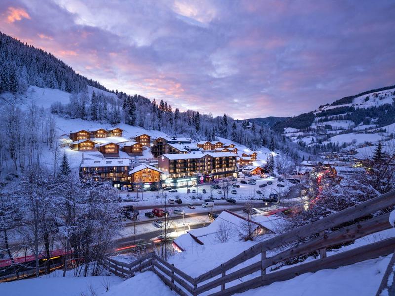 A snowy mountain village with lit chalets at dusk under a purple sky.