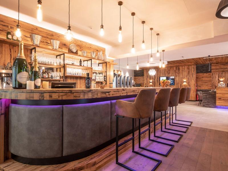 Modern hotel bar area with wooden paneling, hanging light bulbs, and purple-lit counter.
