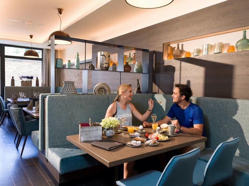 Two people sit at a modern restaurant table with breakfast, talking to each other.
