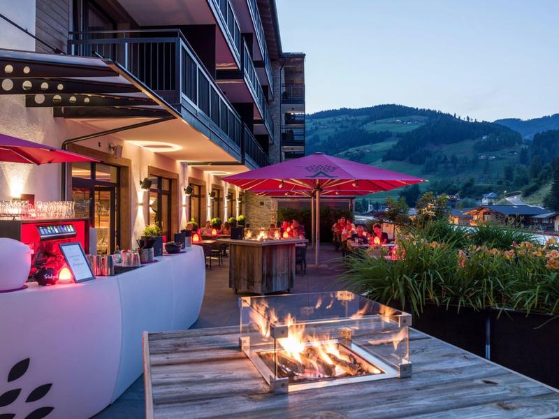 Hotel terrace with fire pit, red umbrellas, and mountain view at dusk.
