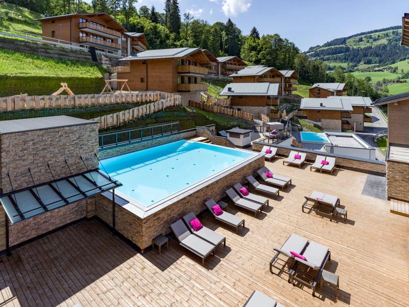 Leisure area with outdoor pool, sun loungers, and mountain chalets in the background on a sunny day.