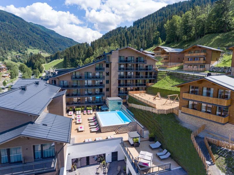 Hotel complex with outdoor pool and sun loungers in a mountainous landscape under blue sky.