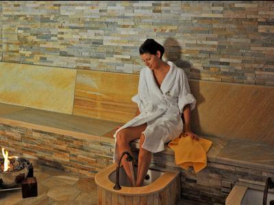 Frau im Bademantel entspannt an einer Fußwanne in einem Wellnessbereich mit Steinwand.