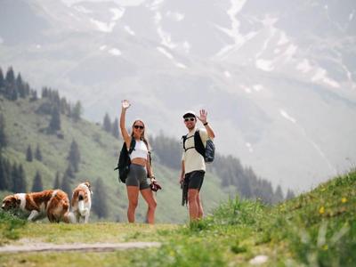Two hikers with backpacks and two dogs wave on a mountain trail.