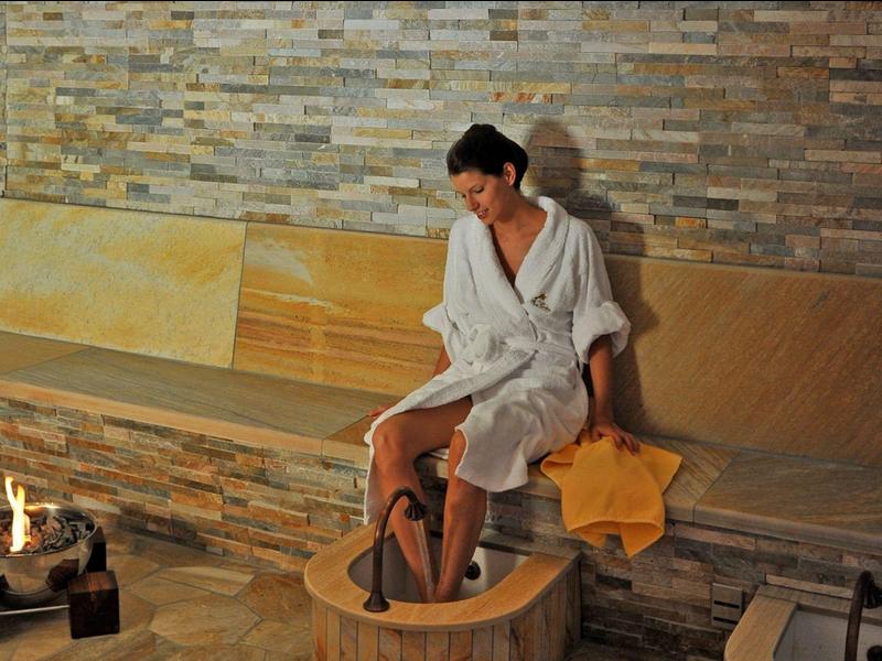 Frau im Bademantel entspannt an einer Fußwanne in einem Wellnessbereich mit Steinwand.