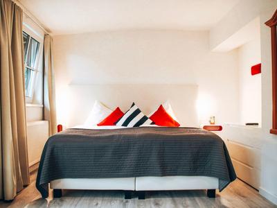 Bright hotel room with a double bed, striped and red pillows, and a large window with beige curtains.