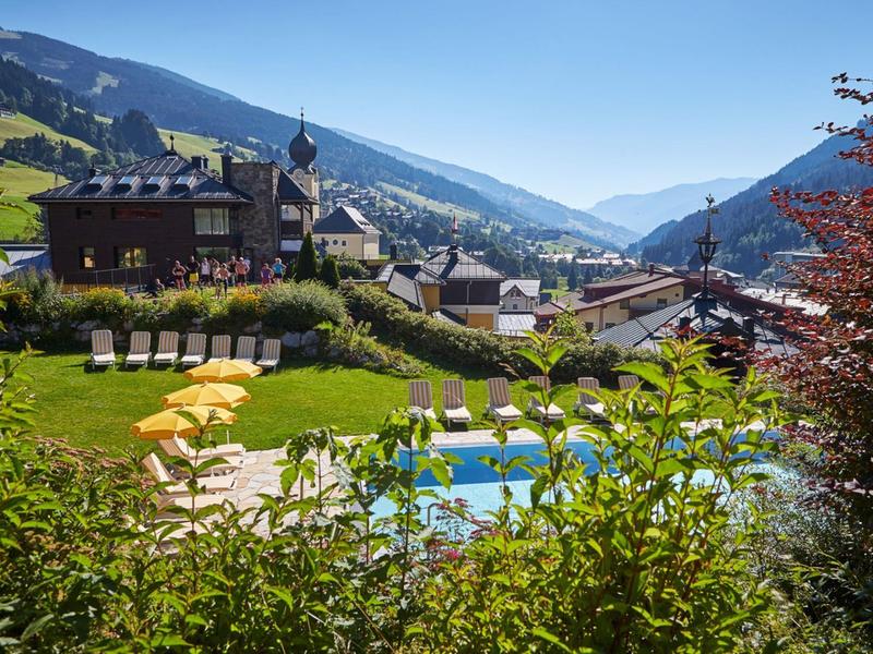 Mountain village scene with pool, lounge chairs, and traditional alpine houses under clear sky.