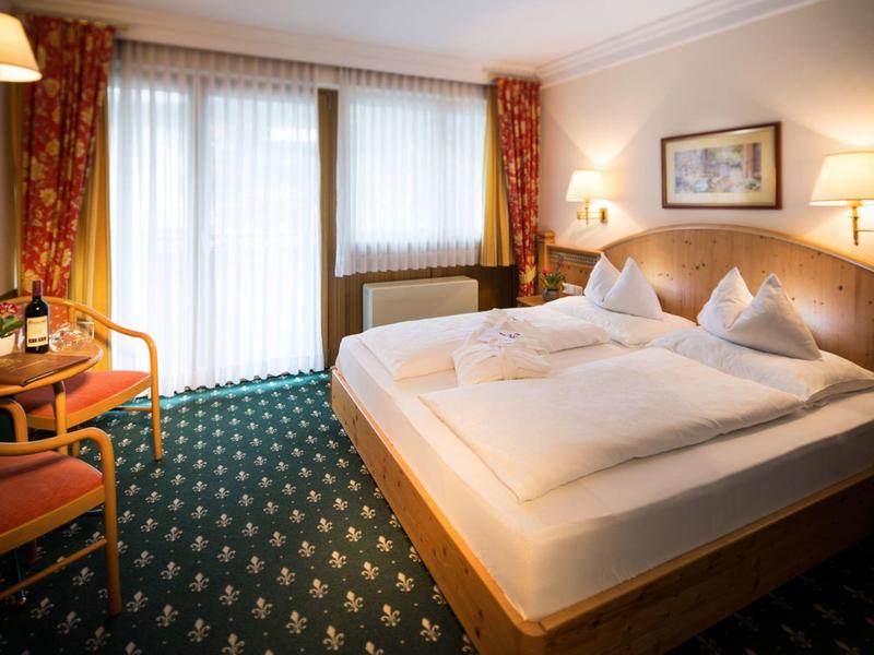 Gemütliches Hotelzimmer mit Doppelbett, zwei roten Stühlen, Teppich und großen Fenstern mit Vorhängen.